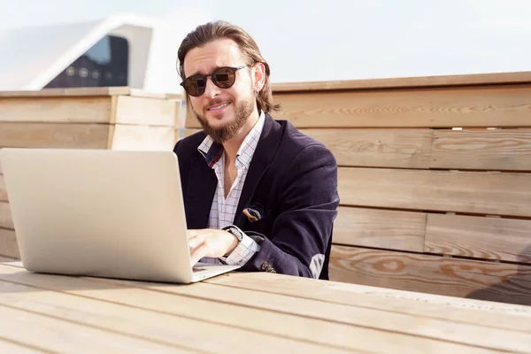 Foto di uomo allegro in occhiali da sole seduto con computer portatile su una panchina di legno in primavera — Foto Stock