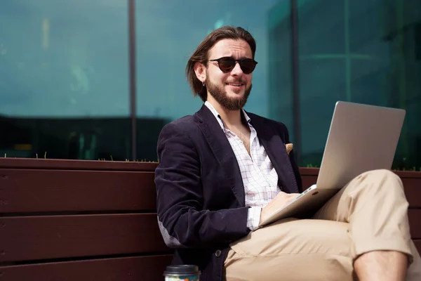 Foto di un uomo freelance sorridente con un computer portatile seduto su una panchina di legno accanto a una parete di vetro — Foto Stock