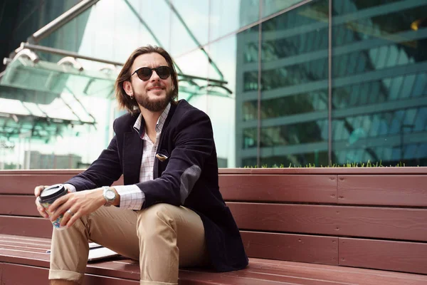 Foto de un hombre moreno con barba mirando hacia un lado con vidrio en la mano sentado en un banco de madera contra el fondo del edificio de vidrio — Foto de Stock