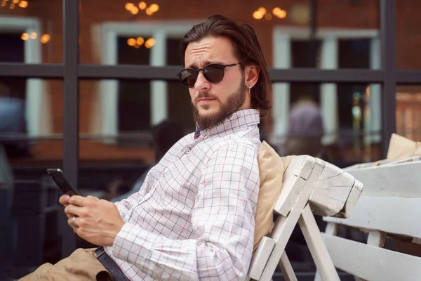 Foto di uomo barbuto in occhiali da sole con il telefono in mano guardando in macchina fotografica mentre seduto in strada caffè in città — Foto Stock