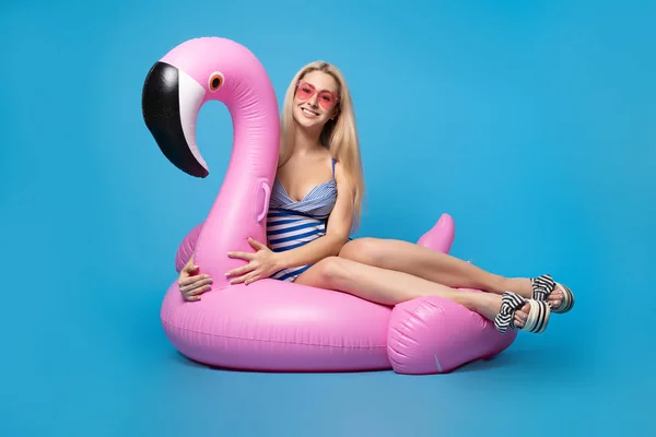 Photo of happy blonde in bathing suit and in pink glasses lying on pink inflatable flamingo on empty blue background — Stock Photo, Image