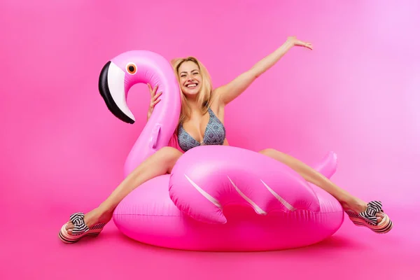 Photo of blonde in bathing suit and pink glasses with raised hand up sitting on inflatable flamingo on empty pink background — Stock Photo, Image