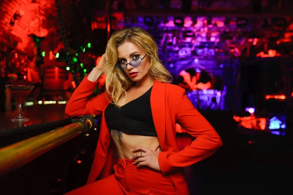 Foto av blond kvinna i röd jacka i svarta glasögon sitter nära bar i nattklubb — Stockfoto