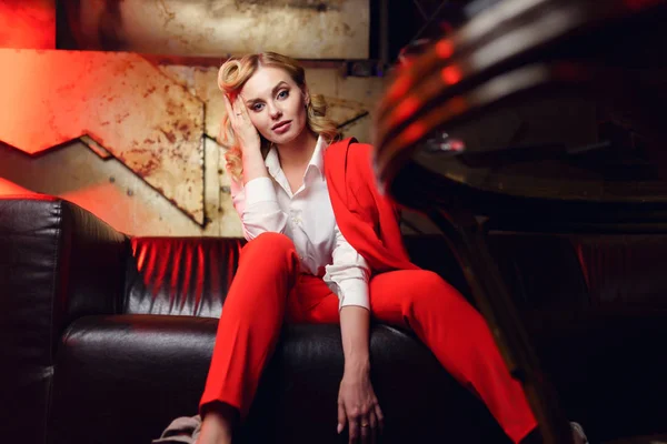 Foto av allvarlig blondin i röd jacka sitter på skinnsoffa — Stockfoto