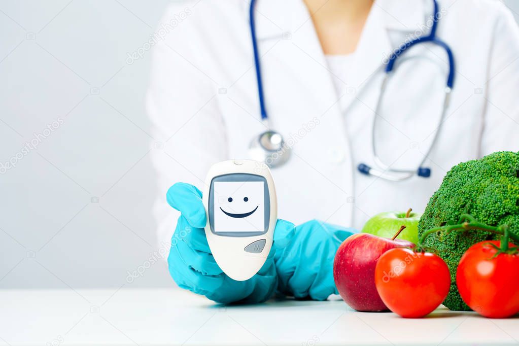 Photo of doctor in lab coat and rubber gloves with glucometer showing smiley at table with vegetables on gray background