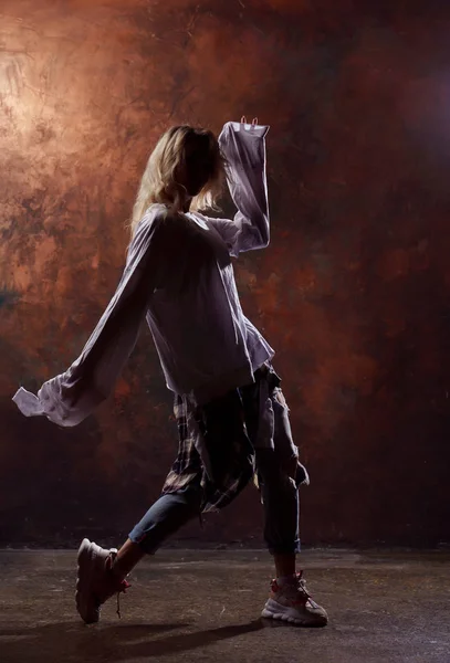 Foto de jovem loira dançando olhando para o lado em jeans rasgados e tênis em fundo marrom escuro — Fotografia de Stock