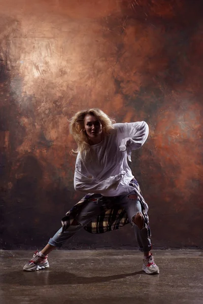 Φωτογραφία του μακριά μαλλιά ξανθιά χορεύτρια κοιτάζοντας την κάμερα σε σκισμένο τζιν και αθλητικά παπούτσια σε σκούρο καφέ φόντο — Φωτογραφία Αρχείου