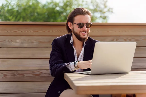 Foto di felice uomo bruno in occhiali da sole seduto con computer portatile su una panchina di legno in primavera — Foto Stock