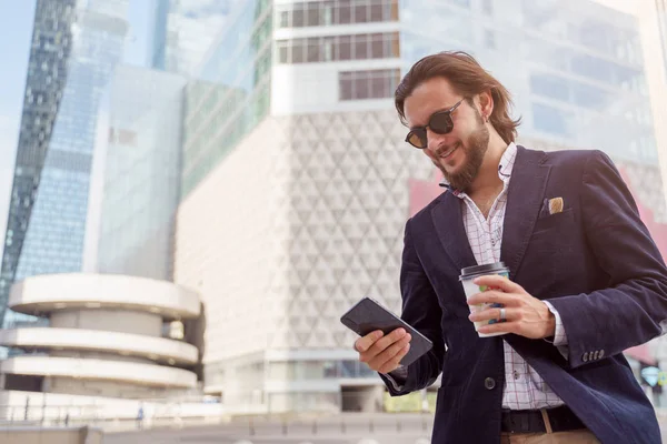 Foto di felice brunetta maschio in occhiali da sole con il telefono in mano mentre passeggia per la città — Foto Stock