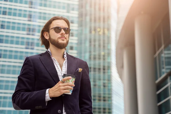 Foto de un hombre moreno con barba mirando hacia un lado con vidrio en la mano en la ciudad contra el fondo de edificios altos — Foto de Stock