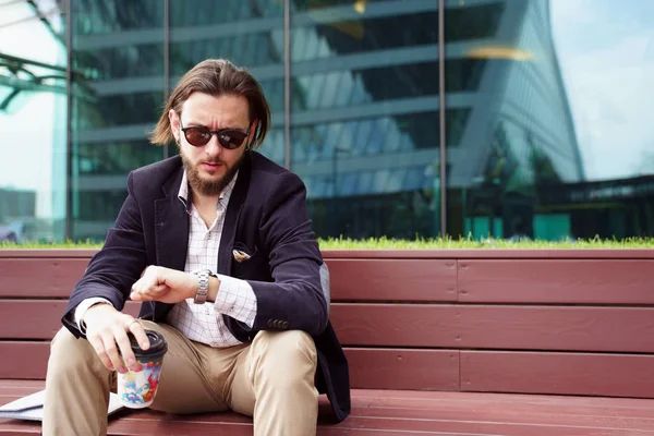 Foto de la elegante morena mirando el reloj en la mano mientras está sentado en un banco de madera contra el fondo del edificio de vidrio —  Fotos de Stock