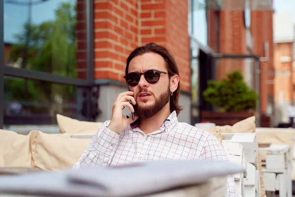 Foto del hombre hablando por teléfono sentado en la cafetería de la calle en la ciudad en la tarde de verano —  Fotos de Stock