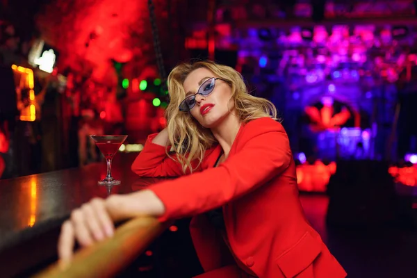 Zdjęcie młodej Blondynki z okularami i czerwonym garniturze siedząc w barze z kieliszkami koktajlowymi w klubie nocnym — Zdjęcie stockowe