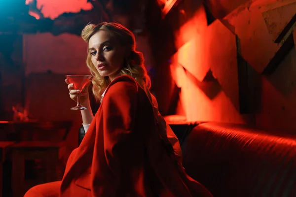 Imagine a tinerei blonde în jachetă roșie uitându-se la camera cu cocktail în mână în clubul de noapte — Fotografie, imagine de stoc