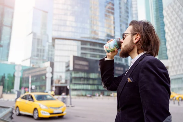Foto a lato di bruna con barba e orecchino in orecchio bere dal vetro in città sullo sfondo di grattacieli, auto gialla — Foto Stock