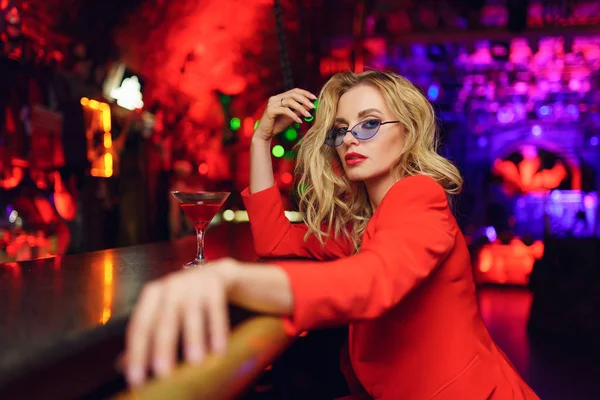 Zdjęcie młodej blond kobiety w okularach i czerwonym garniturze siedzącej w barze z kieliszkami koktajlowymi w klubie nocnym — Zdjęcie stockowe