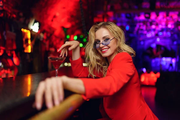 Foto de la rubia feliz con gafas y traje rojo sentado en el bar con copa de cóctel en la discoteca — Foto de Stock