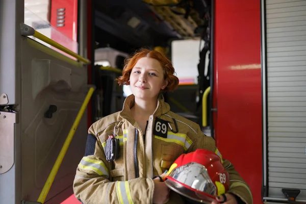 Foto de mujer joven bombero con casco en las manos contra el telón de fondo de camión de bomberos — Foto de Stock