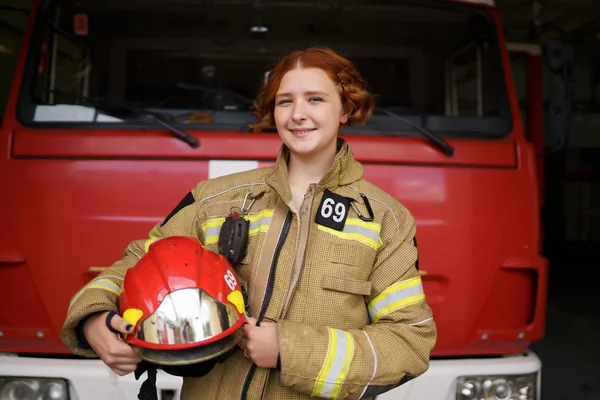 Foto de mujer bombero sonriente con casco en las manos contra el telón de fondo de camión de bomberos — Foto de Stock
