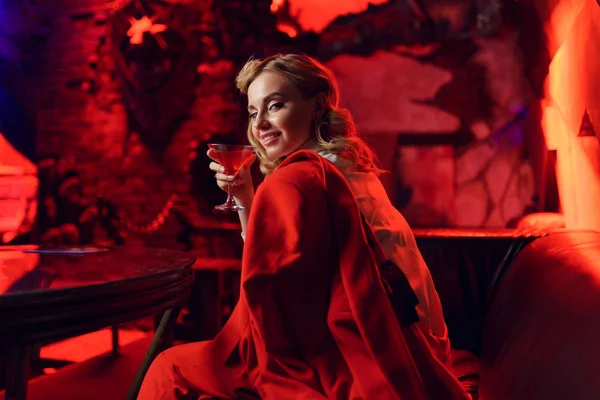 Фото счастливой блондинки в красной куртке, смотрящей в камеру с коктейлем в руке в ночном клубе — стоковое фото