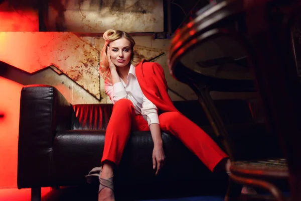 Полнометражное фото молодой блондинки в красной куртке, смотрящей в камеру, сидящую на кожаном диване — стоковое фото