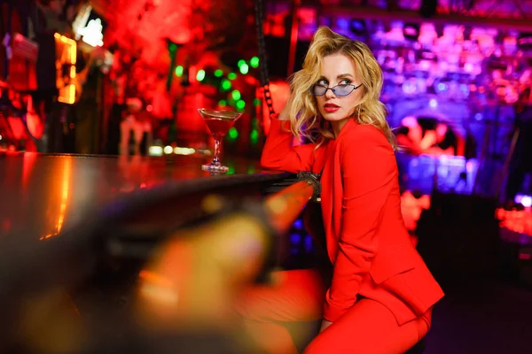 Fotografie a unei femei blonde serioase cu ochelari și costum roșu care se uită la o parte așezată la bar cu sticlă de cocktail în clubul de noapte pe fundal roșu — Fotografie, imagine de stoc