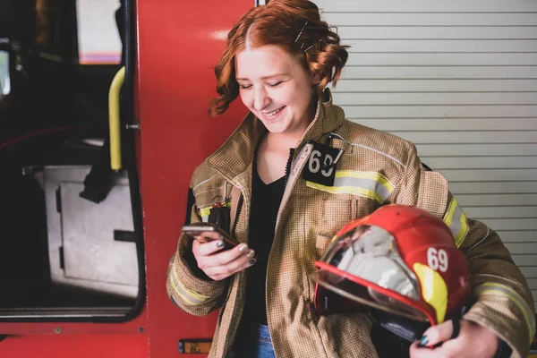 Фото рыжей женщины-пожарного с телефоном в руках на фоне пожарной машины — стоковое фото