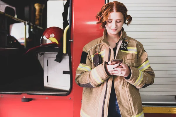 Foto einer jungen Feuerwehrfrau mit Handy in der Hand vor dem Hintergrund eines Feuerwehrfahrzeugs — Stockfoto