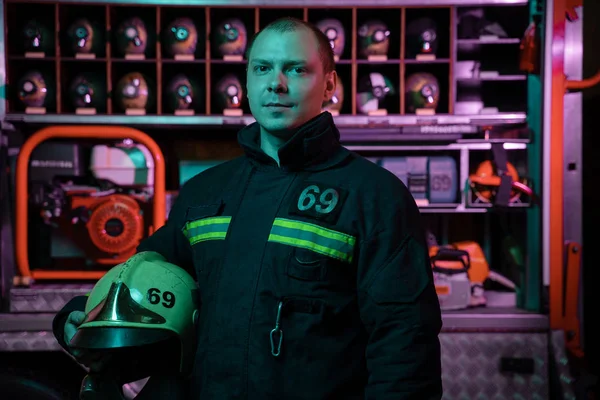 Portrét vážného hasiče, který se dívá na kameru s přilbou v ruce v ohnivé uniformě poblíž hasičového vozu — Stock fotografie