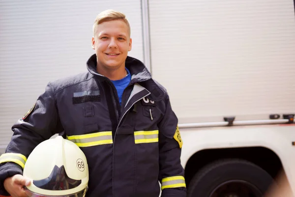 Ritratto di pompiere felice con casco in mano in piedi vicino camion dei pompieri nel pomeriggio . — Foto Stock