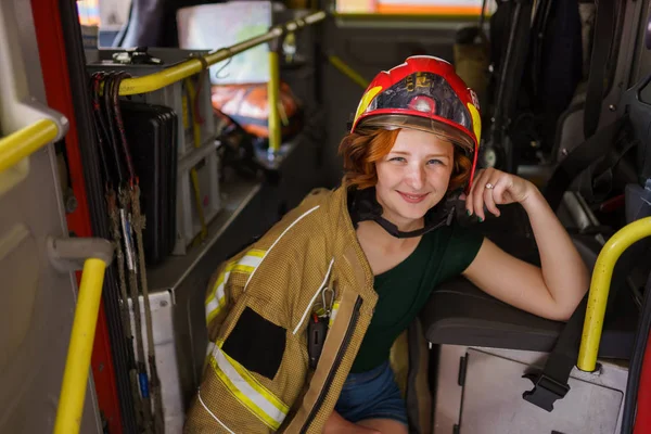 Imagen de la sonriente bombera de jengibre mirando en cámara sentada en cabina del camión de bomberos — Foto de Stock