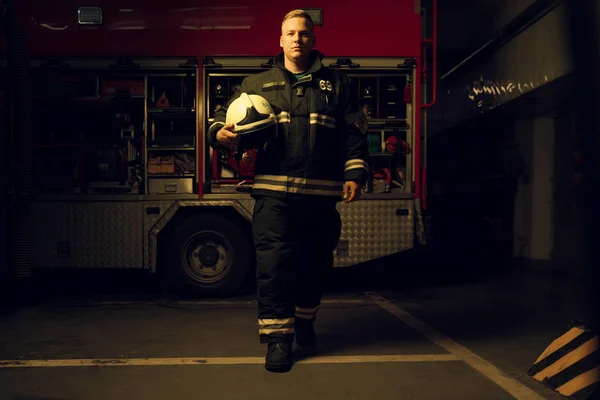 Full-length photo of young firefighter man with helmet in his hand looking into camera while