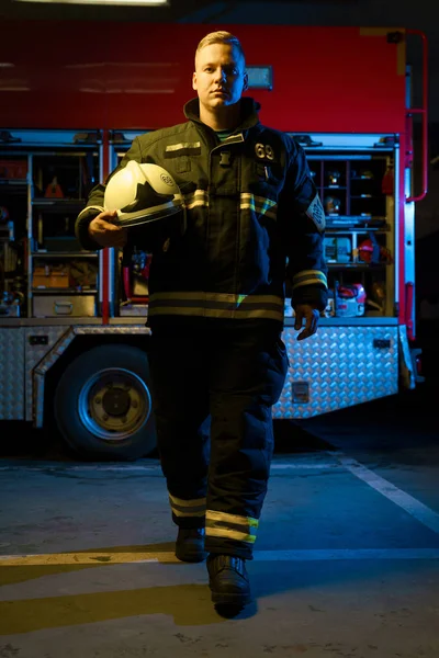 Full-length photo of young firefighter with helmet in his hand looking into camera while standing near fire truck — Stock Photo, Image