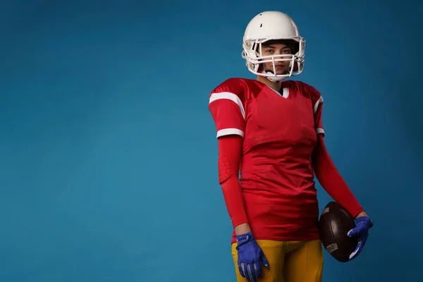 Image of american female football player in white helmet, sportswear and ball in her hand at empty blue background