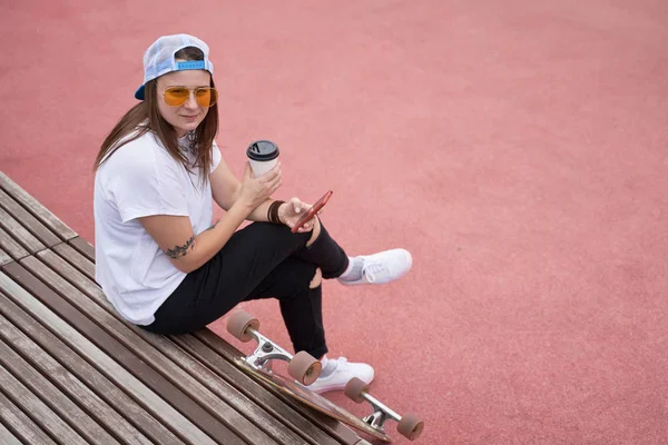 Fotka brunetky se sklenicí a telefonem v dlaních sedícího na dřevěné lavici poblíž skateboard v letním dni — Stock fotografie