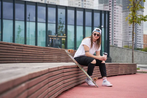 Imagem de esportista morena em óculos de sol com vidro nas mãos sentado em banco de madeira perto de skate no dia de verão — Fotografia de Stock