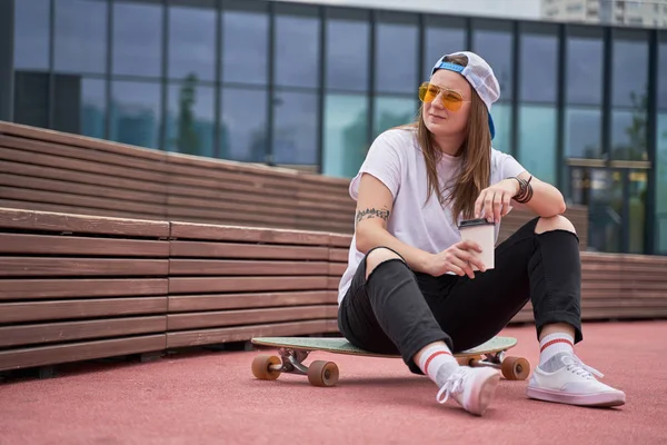 Zdjęcie sportowej Brunetki w okularach w ręce siedzi na deskorolce na boisku sportowym w pobliżu drewnianej ławki w letni dzień — Zdjęcie stockowe