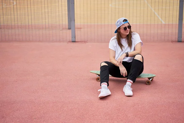 Portrait of athletic brunette in sunglasses sitting on skateboard on playground on summer day — Stock Photo, Image