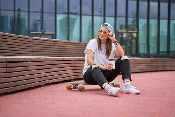 Retrato de esportes morena em óculos de sol em suas mãos sentado no skate no campo esportivo perto de banco de madeira no dia de verão — Fotografia de Stock