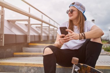 Şehirde merdivenlerde oturan elinde telefon ile güneş gözlüğü esmer kaykaycı Fotoğrafı