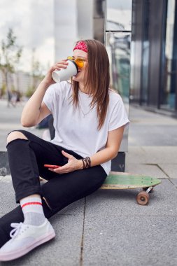 Camdan içme güneş gözlüğü esmer Fotoğraf, elinde telefon şehirde modern binaların arka plan karşı kaykay üzerinde oturan