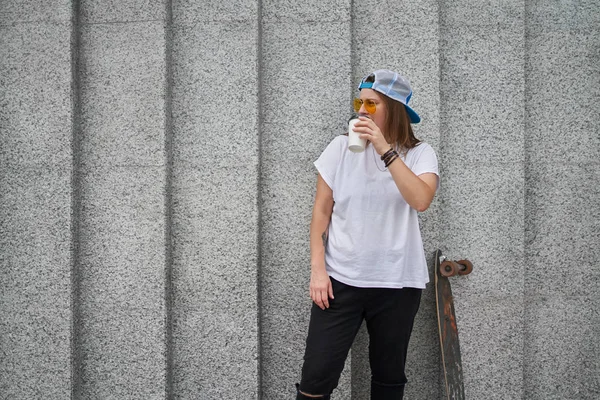 Fotografie mladého sportovce se sklenkou kávy, stojící se skateboard na šedé žulové stěně — Stock fotografie