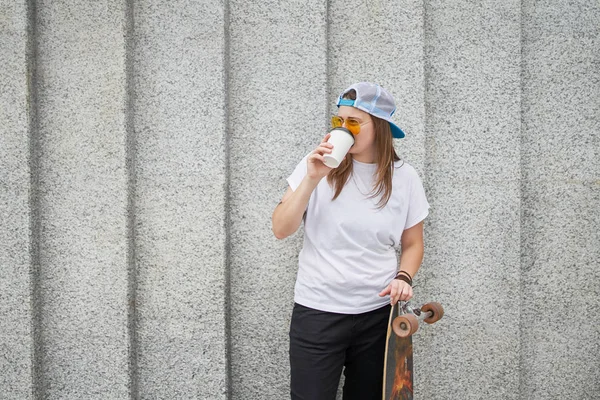 Imagem de jovem atleta feminina com vidro de café, de pé com skate contra parede de granito cinza — Fotografia de Stock