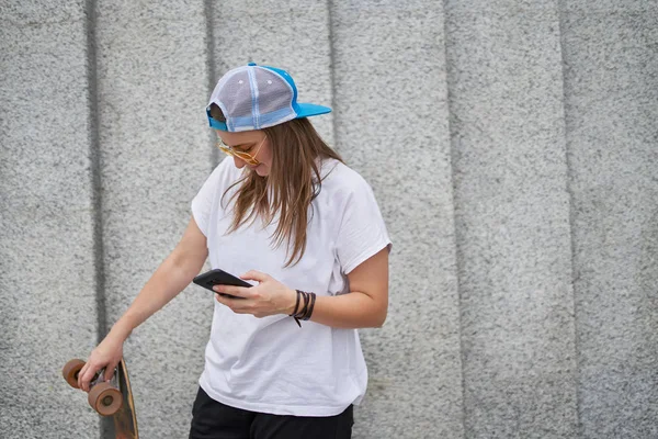 Obraz szczęśliwej kobiecej sportowca w okularze z telefonem w ręku z deskorolka przeciwko szarej ścianie granitu — Zdjęcie stockowe