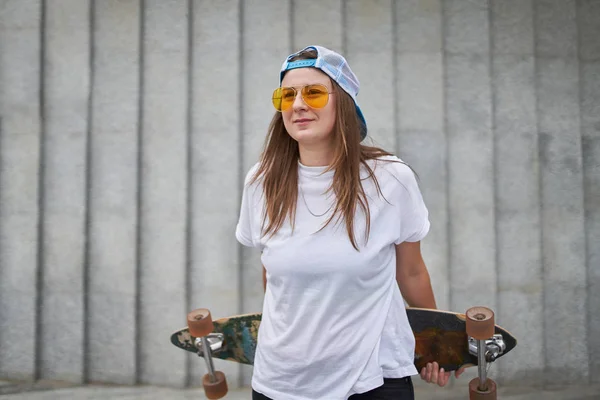 Imagem de atleta feminina feliz em óculos de sol de pé com skate contra a parede de granito cinza — Fotografia de Stock