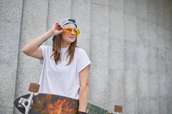 Obrázek Brunetky v slunečních brýlích, stojící se skateboard proti šedé žulové stěně — Stock fotografie