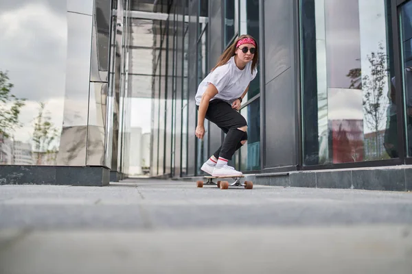 Portret sportowy brunetka jazda na deskorolce na ulicy wśród nowoczesnych budynków — Zdjęcie stockowe