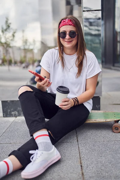 Foto de morena em óculos de sol com telefone e vidro em suas mãos sentadas no skate no fundo de edifícios modernos na cidade — Fotografia de Stock