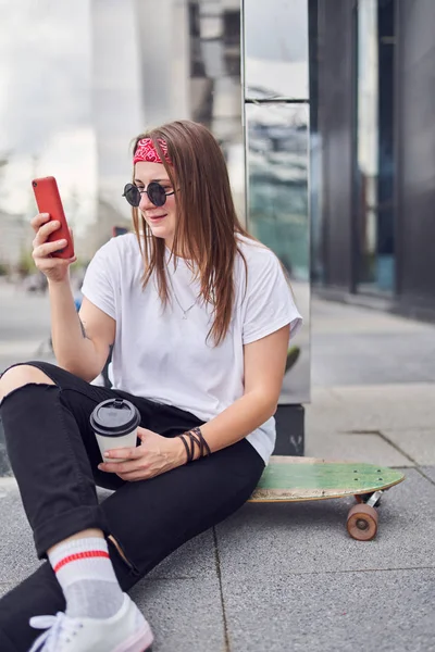 Şehirde modern binaların arka planda kaykay üzerinde oturan elinde telefon ile güneş gözlüğü esmer Fotoğraf