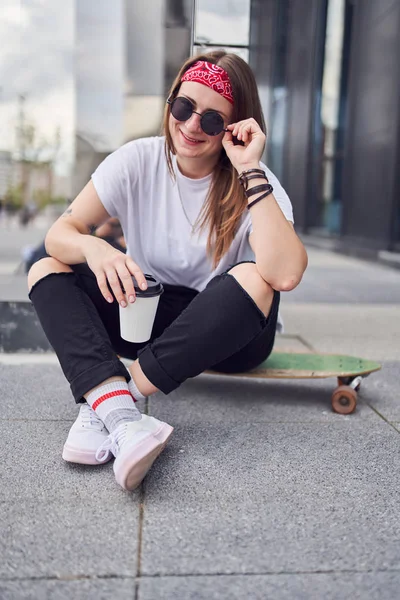 Beeld van jonge brunette vrouw in zonnebrillen kijken naar camera met glas in haar handen zittend op skateboard op achtergrond van moderne gebouwen in de stad — Stockfoto